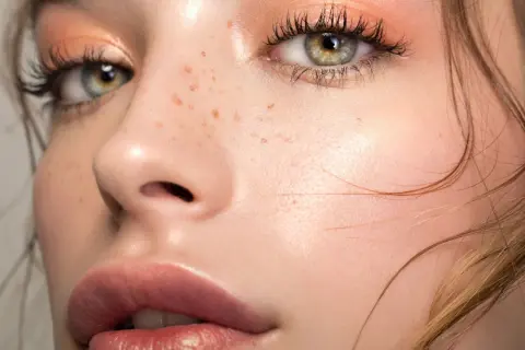 Occhi verdi con trucco nude, ciglia lunghe e labbra piene. La pelle è luminosa con lentiggini naturali.