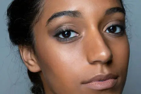 Primo piano di una donna con un intenso trucco occhi nero, labbra nude e pelle luminosa.
