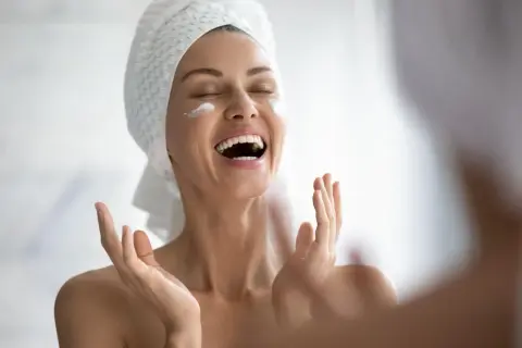 Donna con asciugamano in testa applica crema sul viso, gioia per il trattamento pelle grassa.