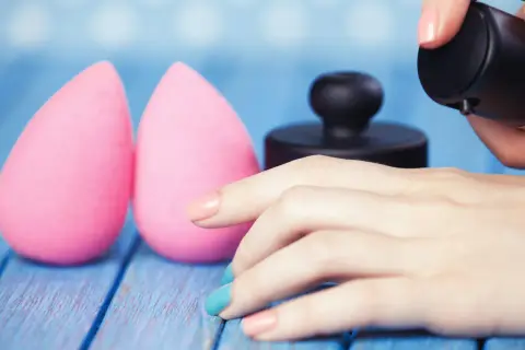 Due spugnette rosa e una mano con smalto colorato che applica fondotinta. Impara come lavare le beauty blender per mantenerle perfette.