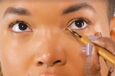 Occhi illuminati da un trucco oro e marrone scintillante, con eyeliner dorato applicato con precisione.