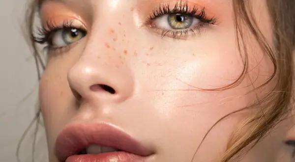Occhi verdi con trucco nude, ciglia lunghe e labbra piene. La pelle è luminosa con lentiggini naturali.