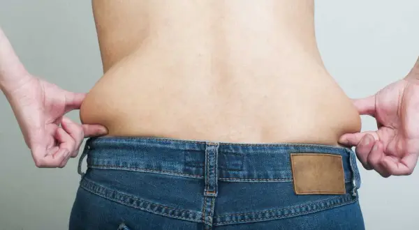 Mani che pizzicano la pelle sui fianchi, evidenziando accumuli di grasso sopra i jeans.