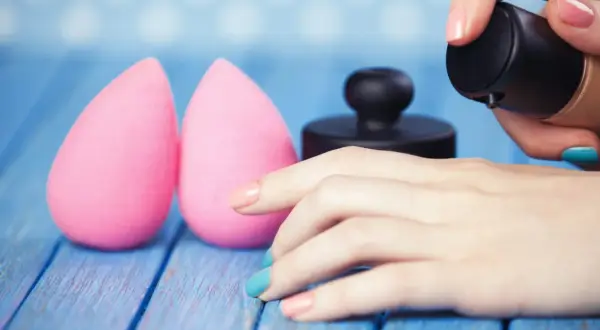 Due spugnette rosa e una mano con smalto colorato che applica fondotinta. Impara come lavare le beauty blender per mantenerle perfette.