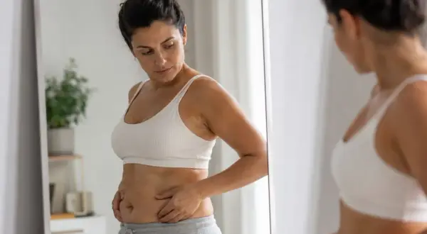Donna si guarda allo specchio, preoccupata per la pancia e i fianchi, pensando a trattamenti estetici per ridurre queste zone.