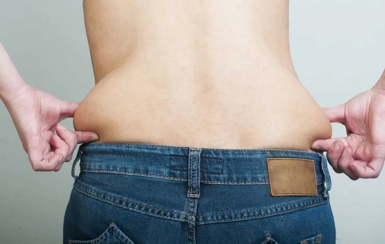 Mani che pizzicano la pelle sui fianchi, evidenziando accumuli di grasso sopra i jeans.