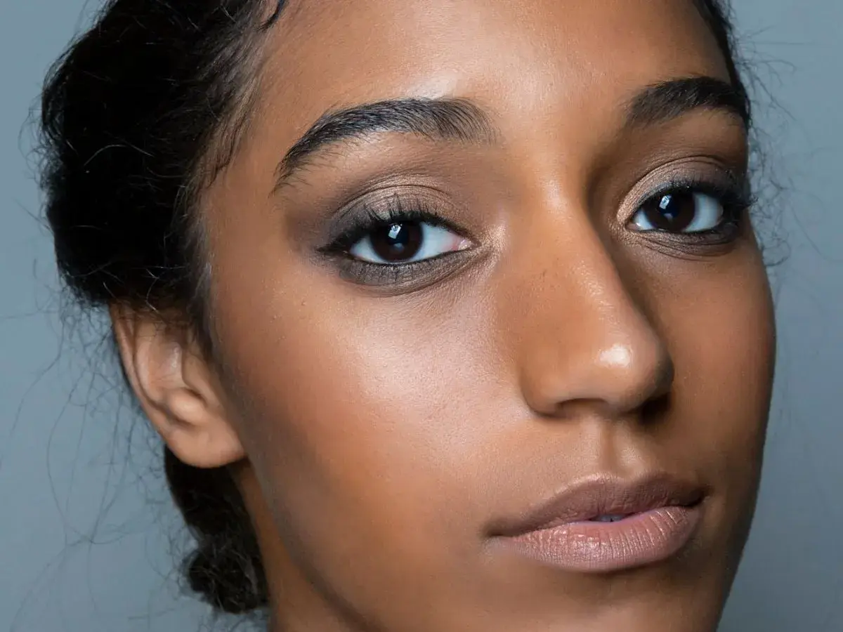 Primo piano di una donna con un intenso trucco occhi nero, labbra nude e pelle luminosa.