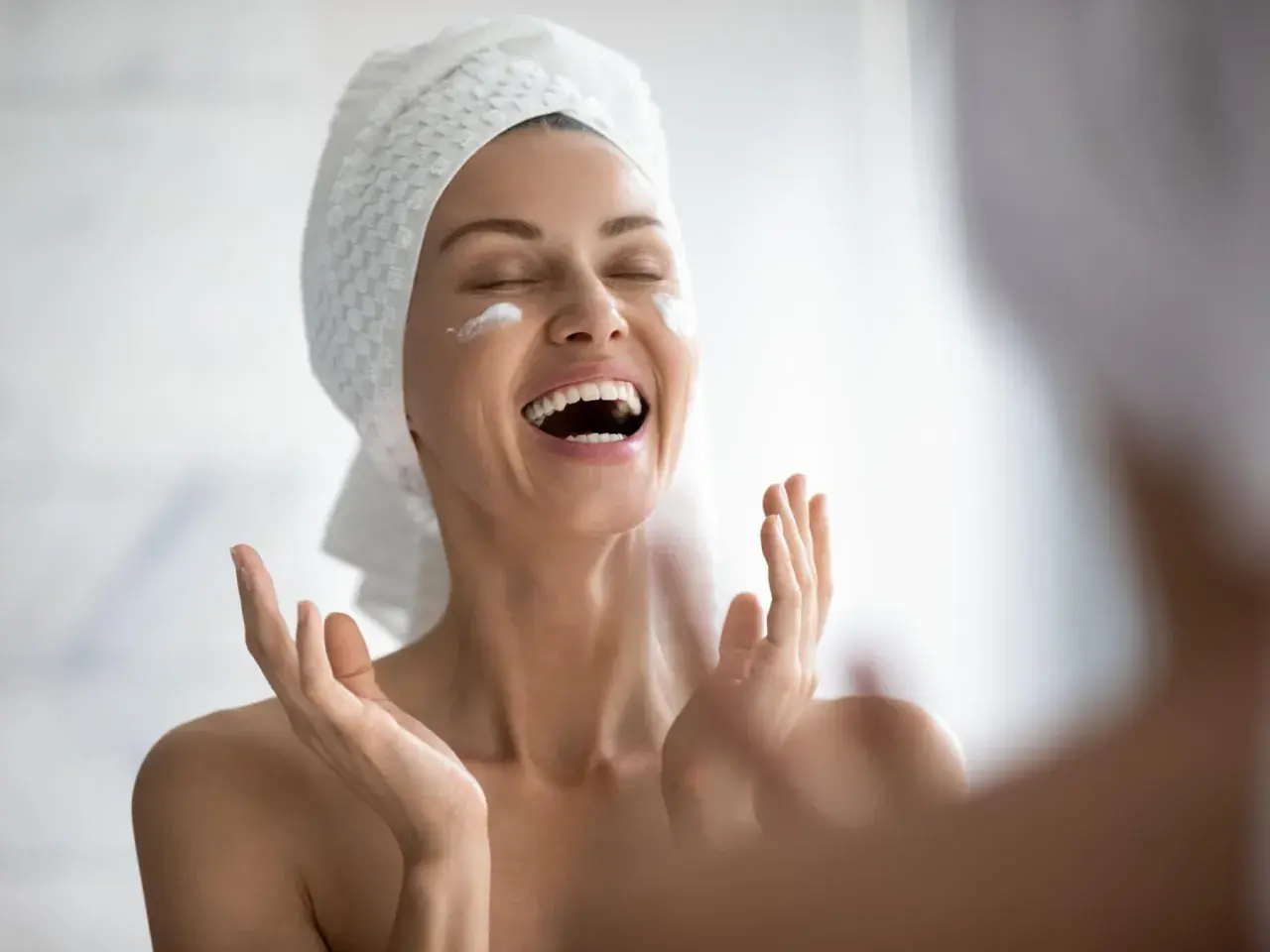 Donna con asciugamano in testa applica crema sul viso, gioia per il trattamento pelle grassa.