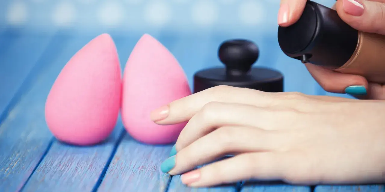 Due spugnette rosa e una mano con smalto colorato che applica fondotinta. Impara come lavare le beauty blender per mantenerle perfette.