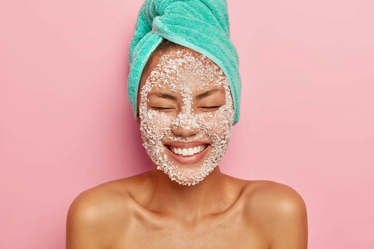 Donna con turbante e scrub sul viso, felice. Lo scrub viso serve a esfoliare e rinnovare la pelle, lasciandola luminosa e liscia.