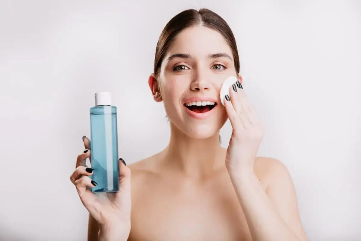 Donna sorridente con tonico blu e dischetto di cotone, pronta per la sua routine di bellezza. Ricorda che il tonico va risciacquato.