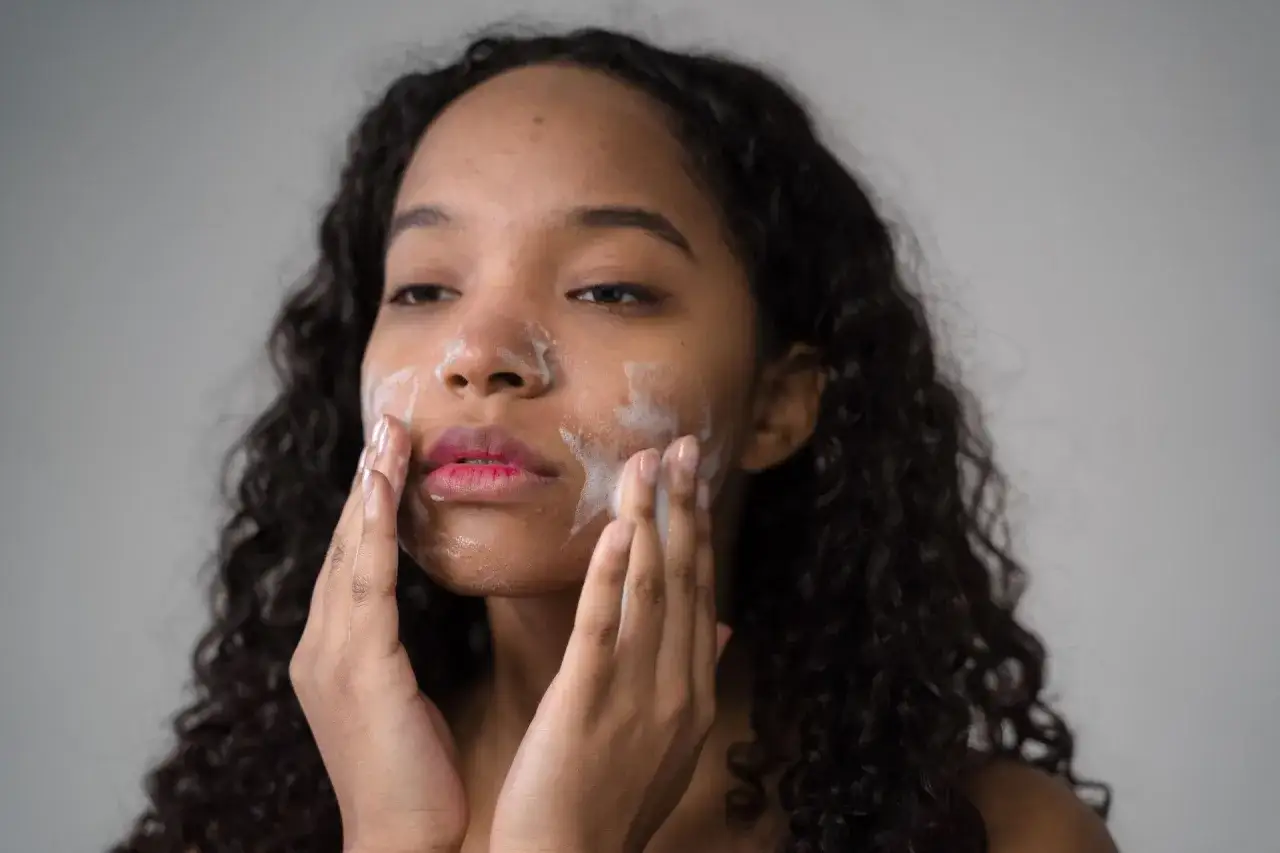 Donna con capelli ricci applica latte detergente viso con le mani, creando una schiuma delicata.