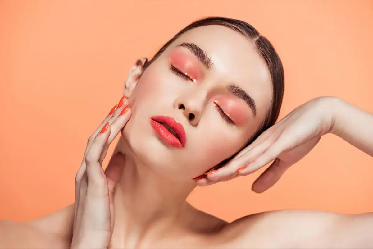 Viso di donna con trucco occhi e labbra sui toni corallo e oro, unghie arancioni. Un'ode alla primavera soft palette.