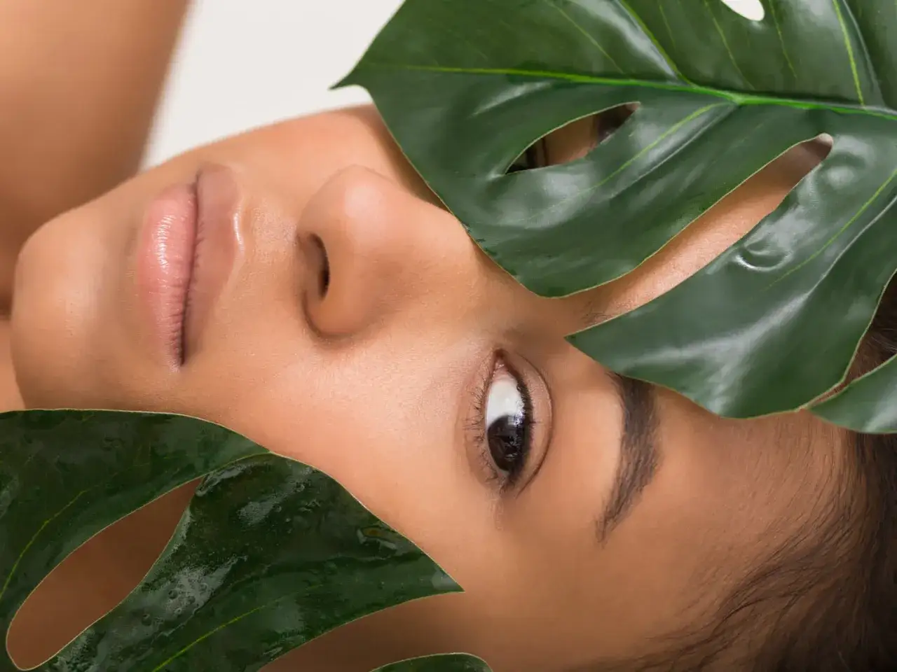 Primo piano del viso di una donna con foglie verdi che incorniciano gli occhi. Un'immagine che evoca benessere e rimedi naturali per la borsa malare.