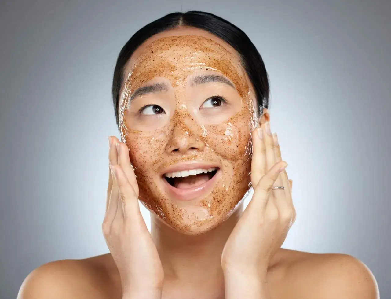 Donna con maschera esfoliante al viso, mani che massaggiano delicatamente. Pelle luminosa e fresca dopo il trattamento.