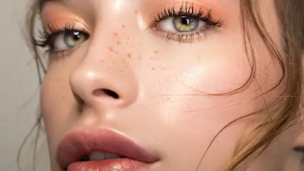 Occhi verdi con trucco nude, ciglia lunghe e labbra piene. La pelle è luminosa con lentiggini naturali.