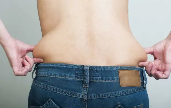 Mani che pizzicano la pelle sui fianchi, evidenziando accumuli di grasso sopra i jeans.