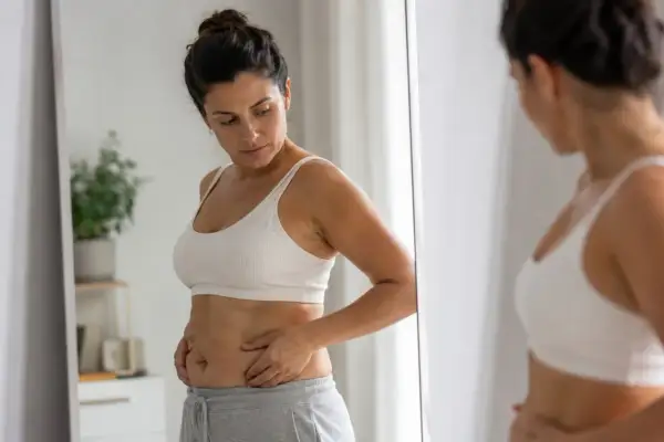 Donna si guarda allo specchio, preoccupata per la pancia e i fianchi, pensando a trattamenti estetici per ridurre queste zone.